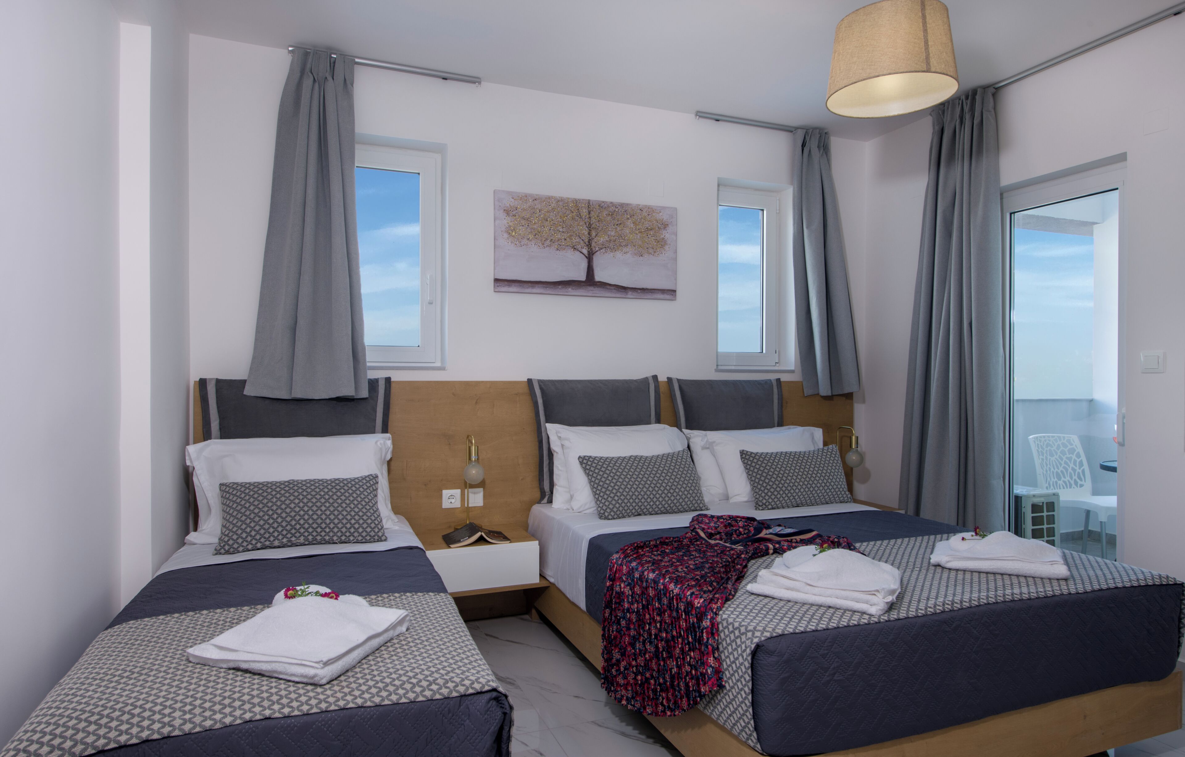 A hotel room with two beds, a bedside table with a lamp, a painting of a tree, and a view through a glass door.