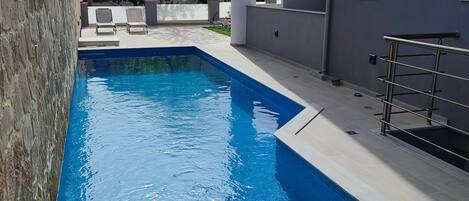 A rectangular swimming pool with clear blue water, surrounded by a stone wall and a modern building with a flat roof.