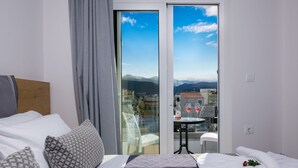 A hotel room with a bed, a balcony with a table and chairs, and a view of the city and mountains. - Deluxe City (Chania)