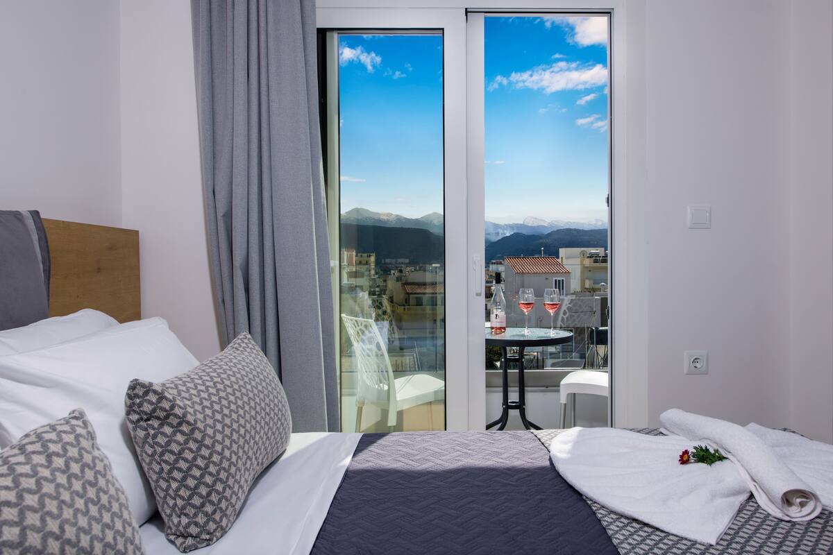 A hotel room with a bed, a balcony with a table and chairs, and a view of the city and mountains.