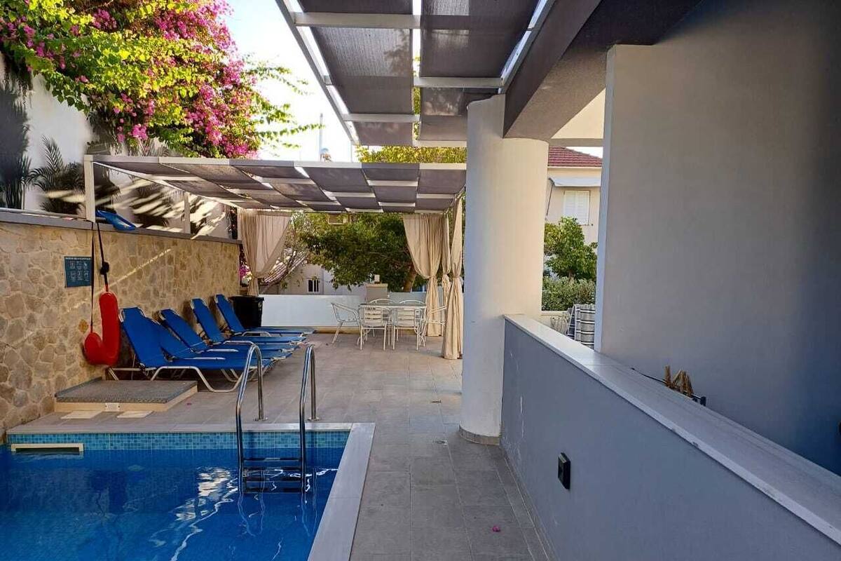 A swimming pool with blue water, surrounded by blue lounge chairs and a covered seating area with a table and chairs.