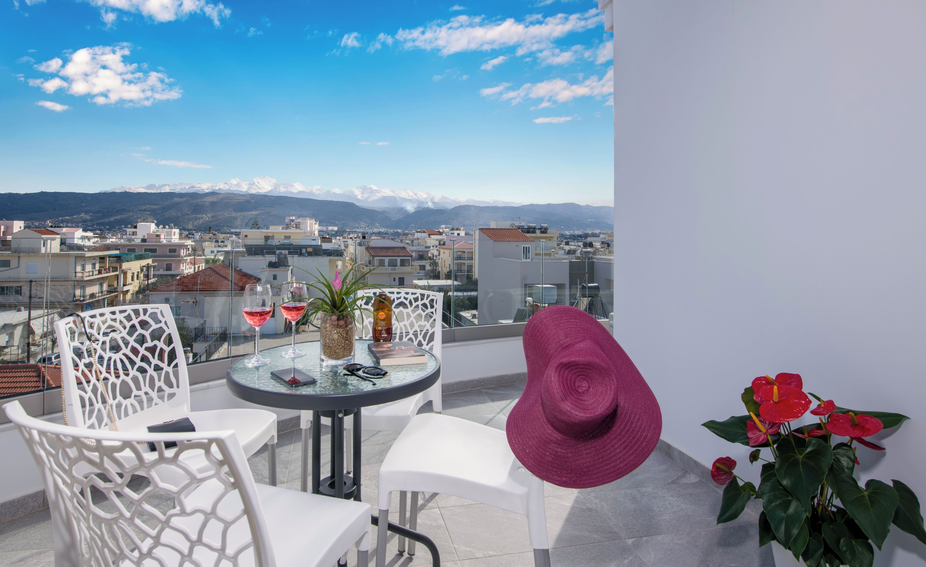 Un balcón con mesa, sillas, una botella y vasos, con vista a una ciudad y montañas al fondo.