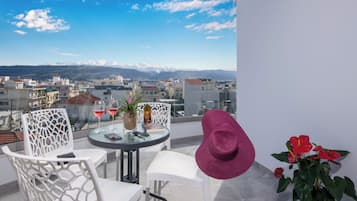Un balcón con mesa, sillas, una botella y vasos, con vista a una ciudad y montañas al fondo.