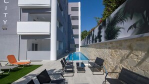 A hotel with a pool, lounge chairs, and a stone wall with tropical plants. - Deluxe City (Chania)