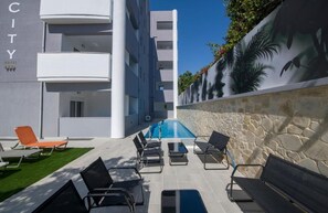 A hotel with a pool, lounge chairs, and a stone wall with tropical plants. - Deluxe City Hotel (Chania)