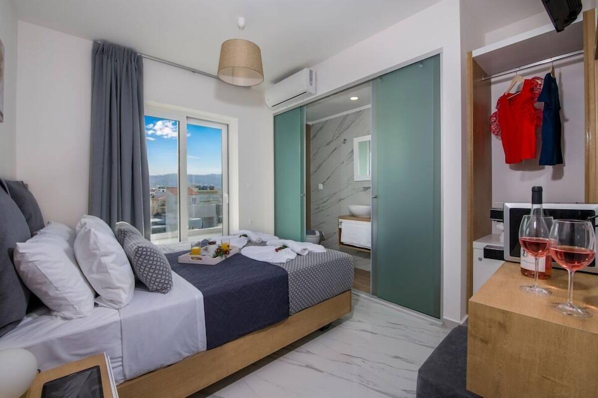 A modern hotel room with a bed, a bedside table with wine glasses, a view of the city, and a sliding door closet.