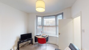 A small, well-lit room with a television, a dining table with chairs, and a large window with blinds. - The Rock Apartments II (London)