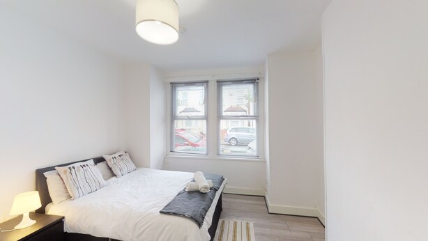 A bedroom with a bed, nightstand, and a window with blinds. - The Rock Apartments II (London)