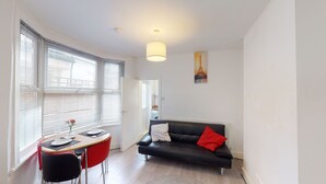 A modern living room with a black leather sofa, a dining table with chairs, and large windows with blinds. - The Rock Apartments II (London)