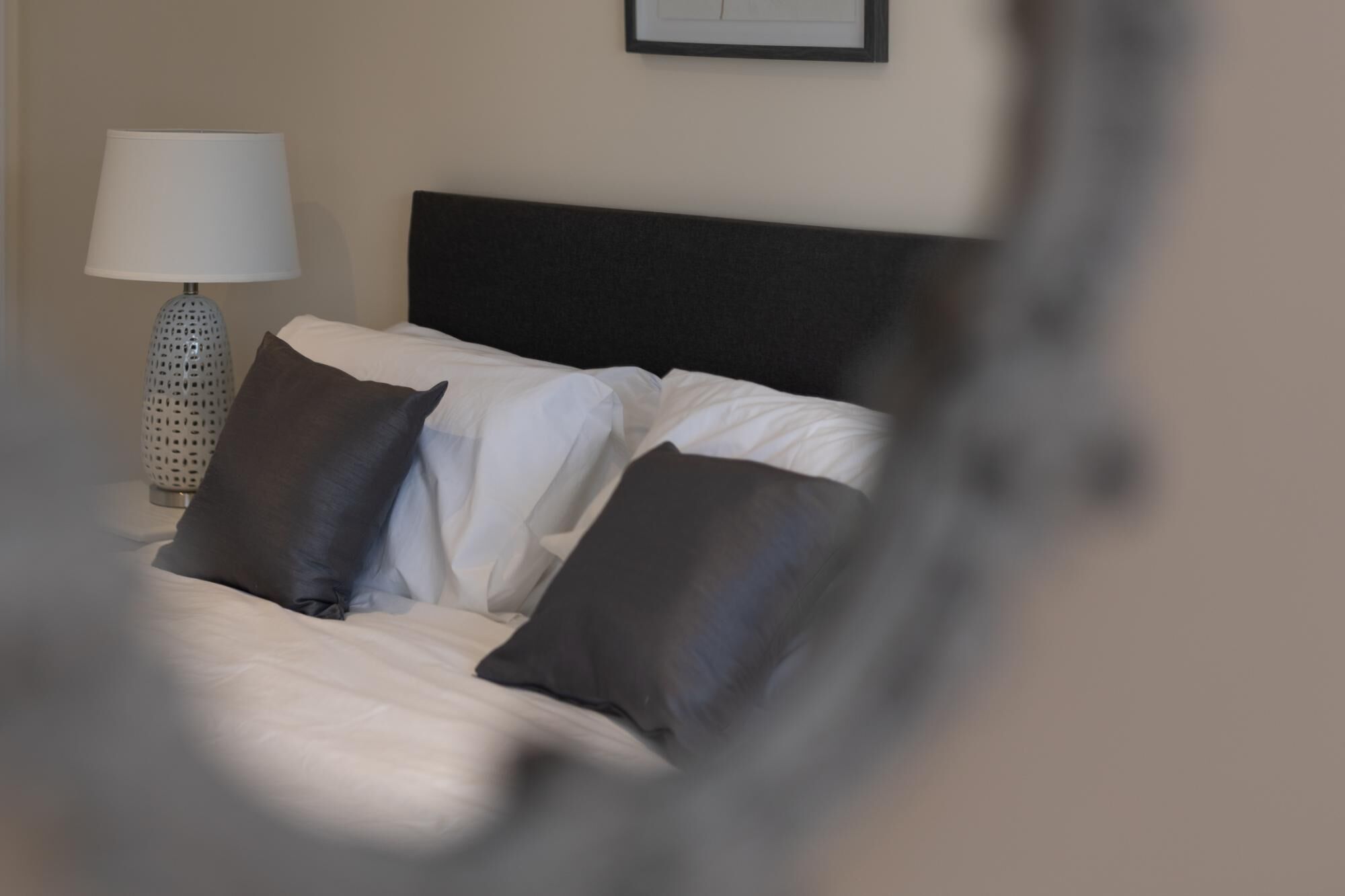 Un lit avec une literie blanche et deux oreillers foncés, une lampe blanche au design ajouré et un tableau encadré accroché au mur.
