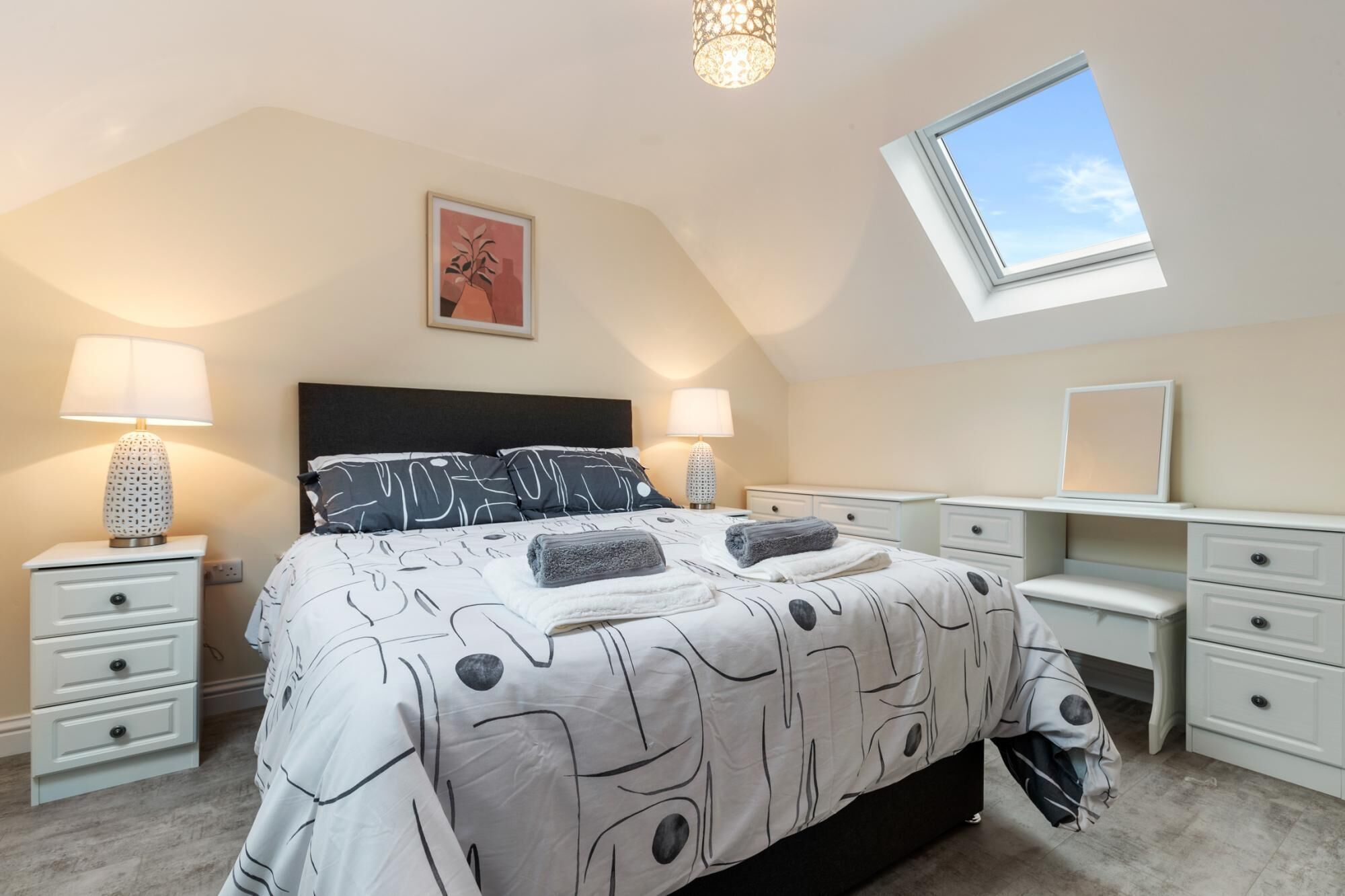 A bedroom with a bed, two bedside tables, a desk, a chair, a window, and a ceiling light fixture.