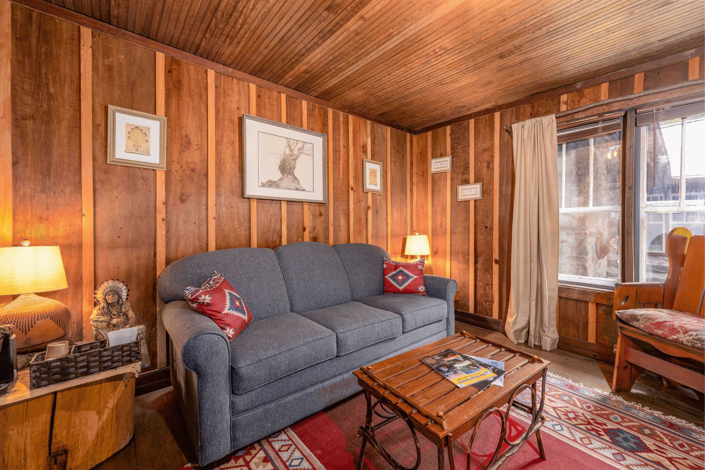 A cozy room with wooden walls, a grey sofa, a wooden coffee table, and a window with curtains.