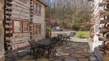 A log cabin with a stone patio, outdoor seating, and a forest backdrop.