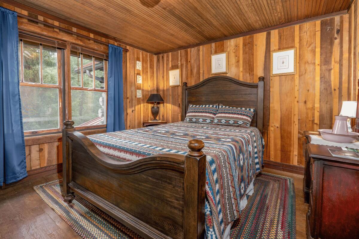A wooden-paneled bedroom with a bed, a nightstand, a lamp, and a window with curtains.