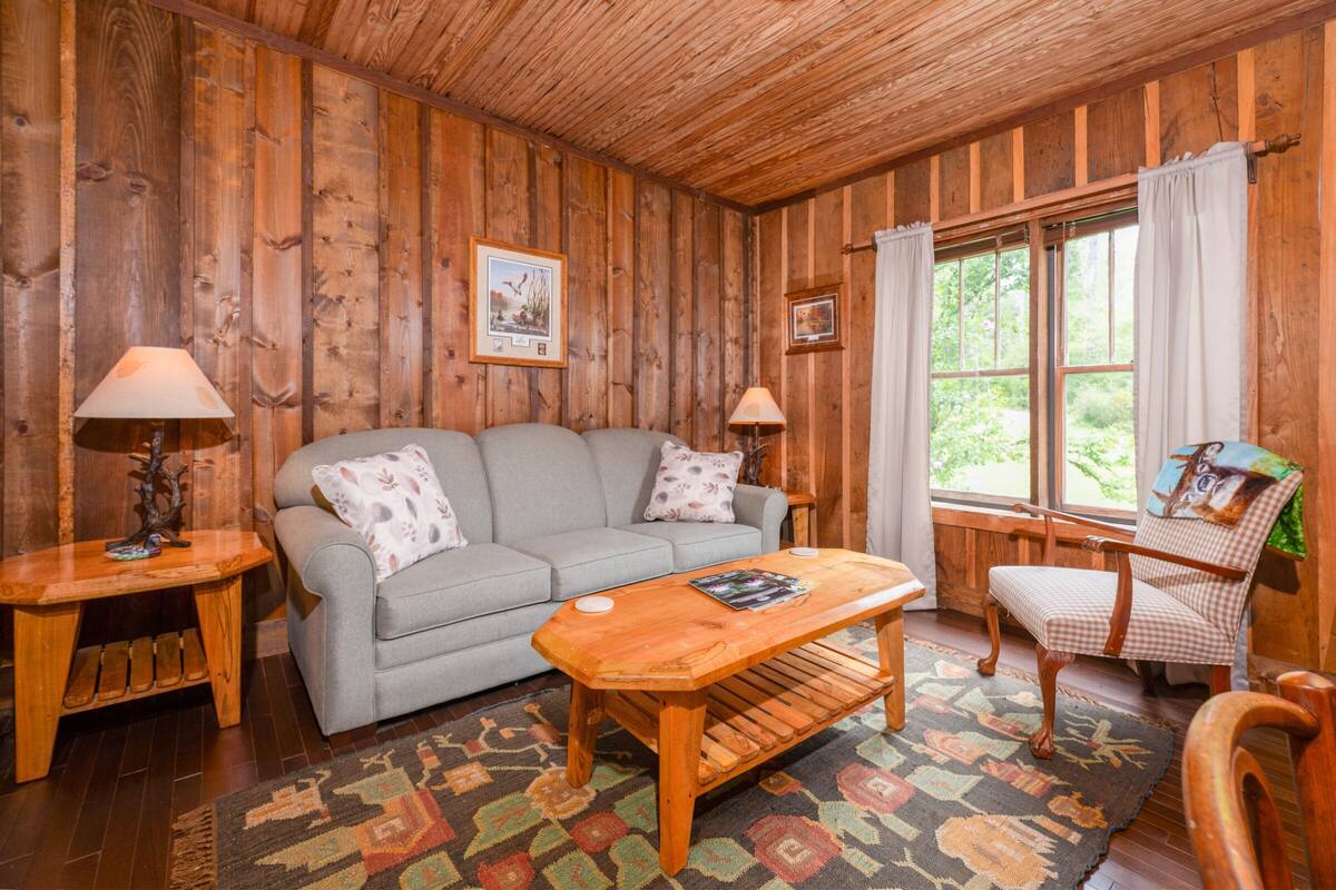 A cozy room with a wooden ceiling, a grey sofa, a wooden coffee table, a patterned rug, and a chair with a checkered cushion.