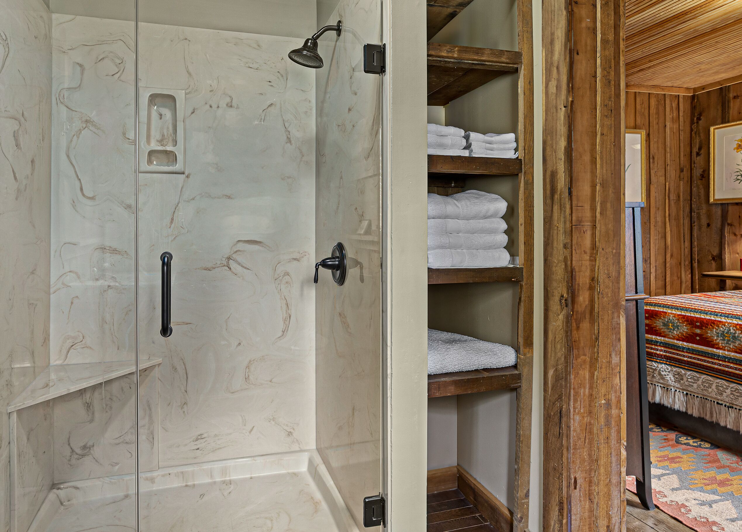 A bathroom with a glass shower enclosure, a wooden shelf with folded towels, and a patterned rug.