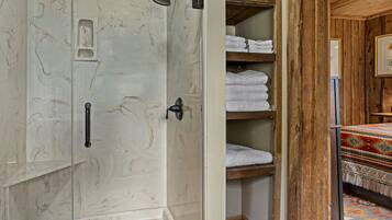 A bathroom with a glass shower enclosure, a wooden shelf with folded towels, and a patterned rug.