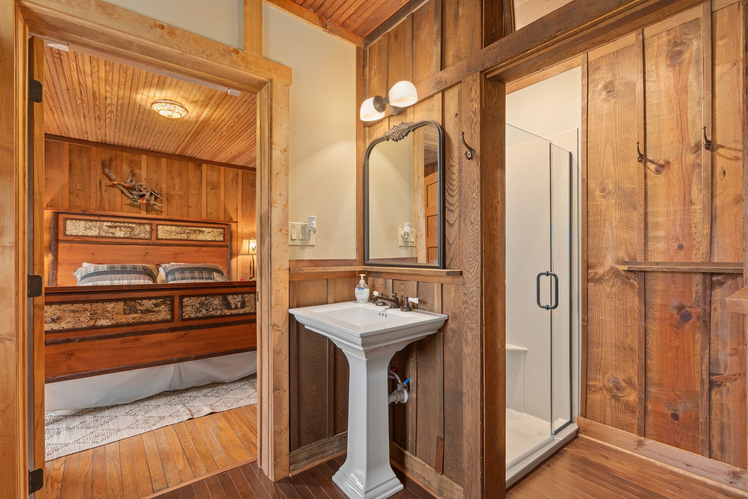 Un baño de madera con lavamanos pedestal, un espejo y una ducha.