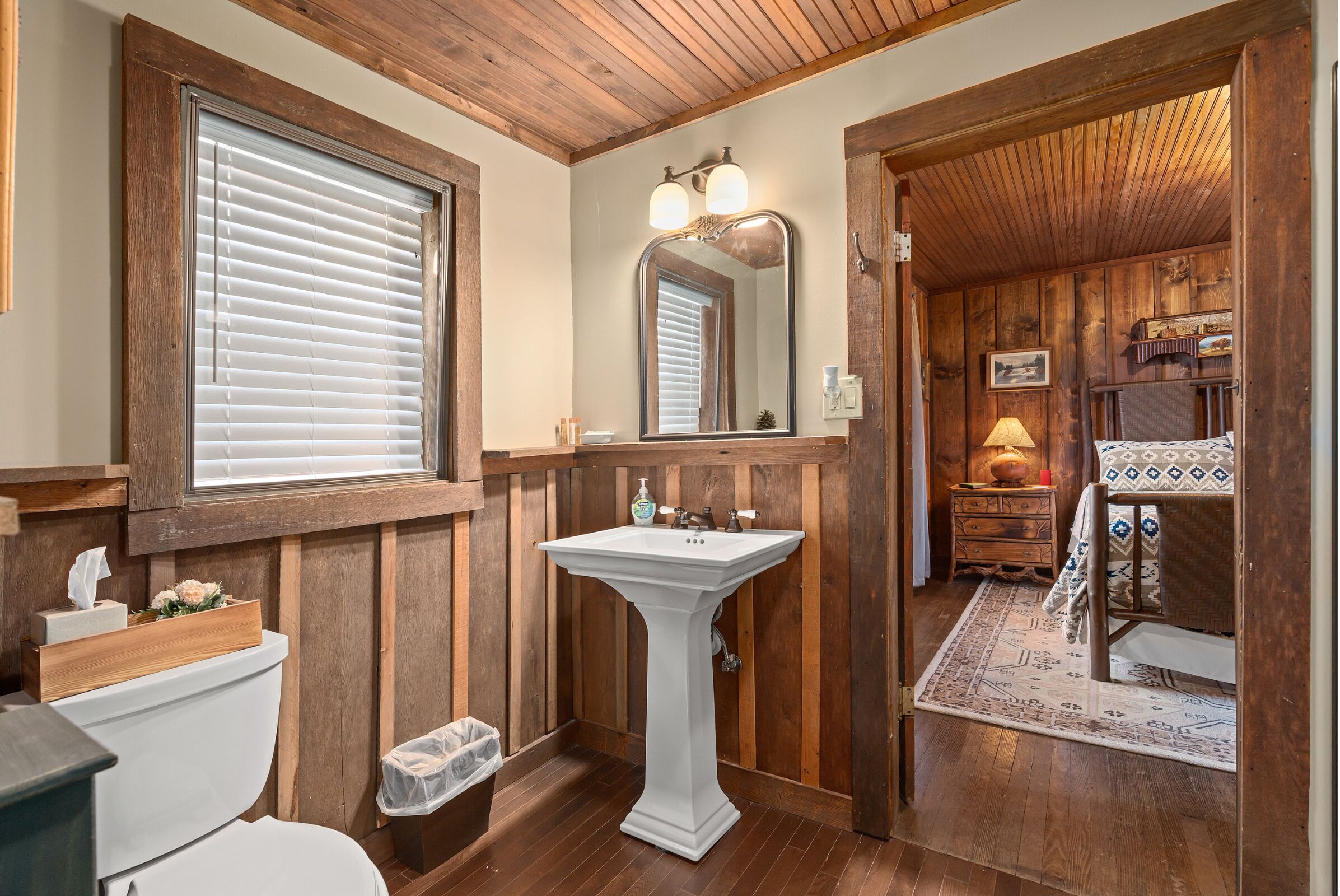 A bathroom with wooden paneling, a pedestal sink, a toilet, and a window with blinds.