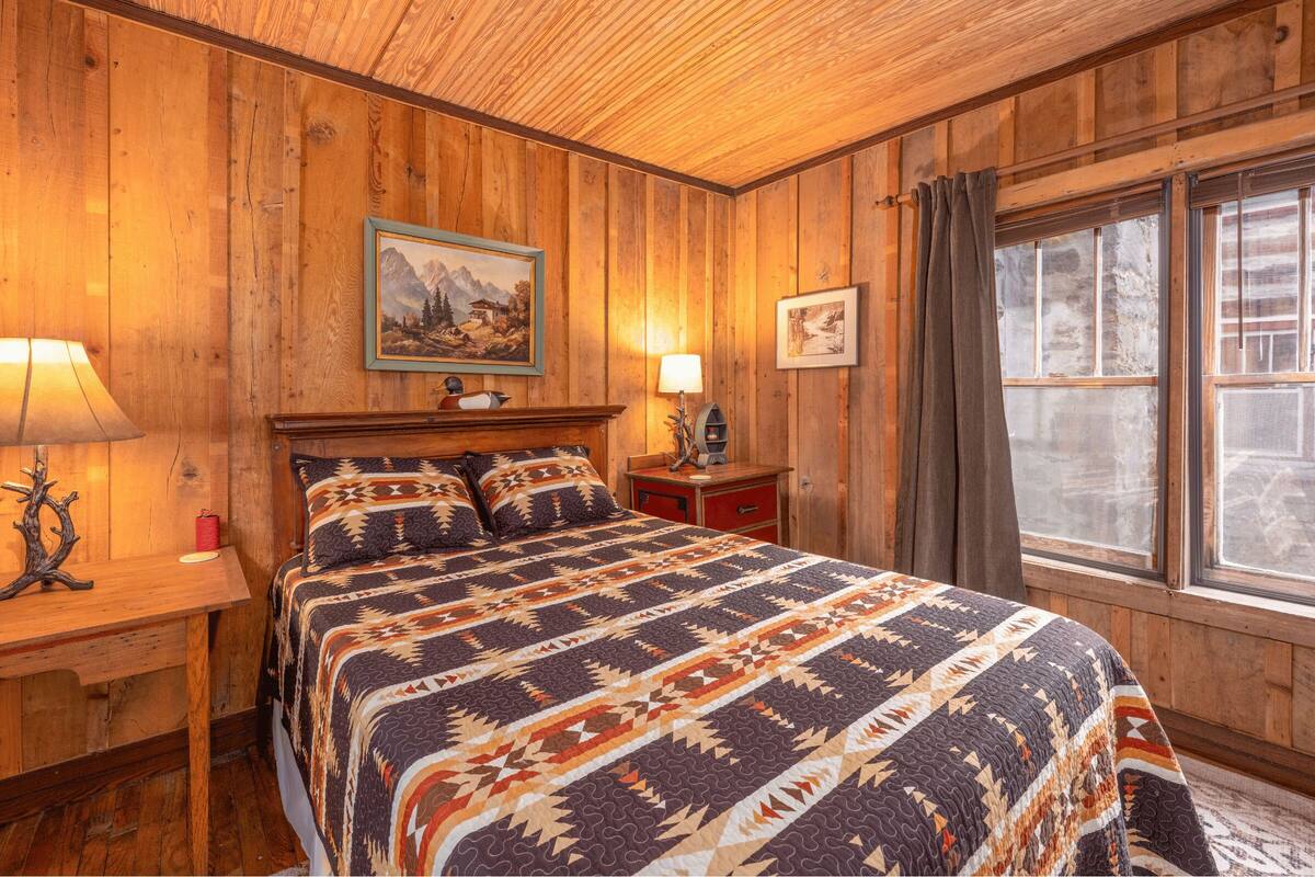 A wooden room with a bed, two bedside tables, a window with curtains, and a painting on the wall.