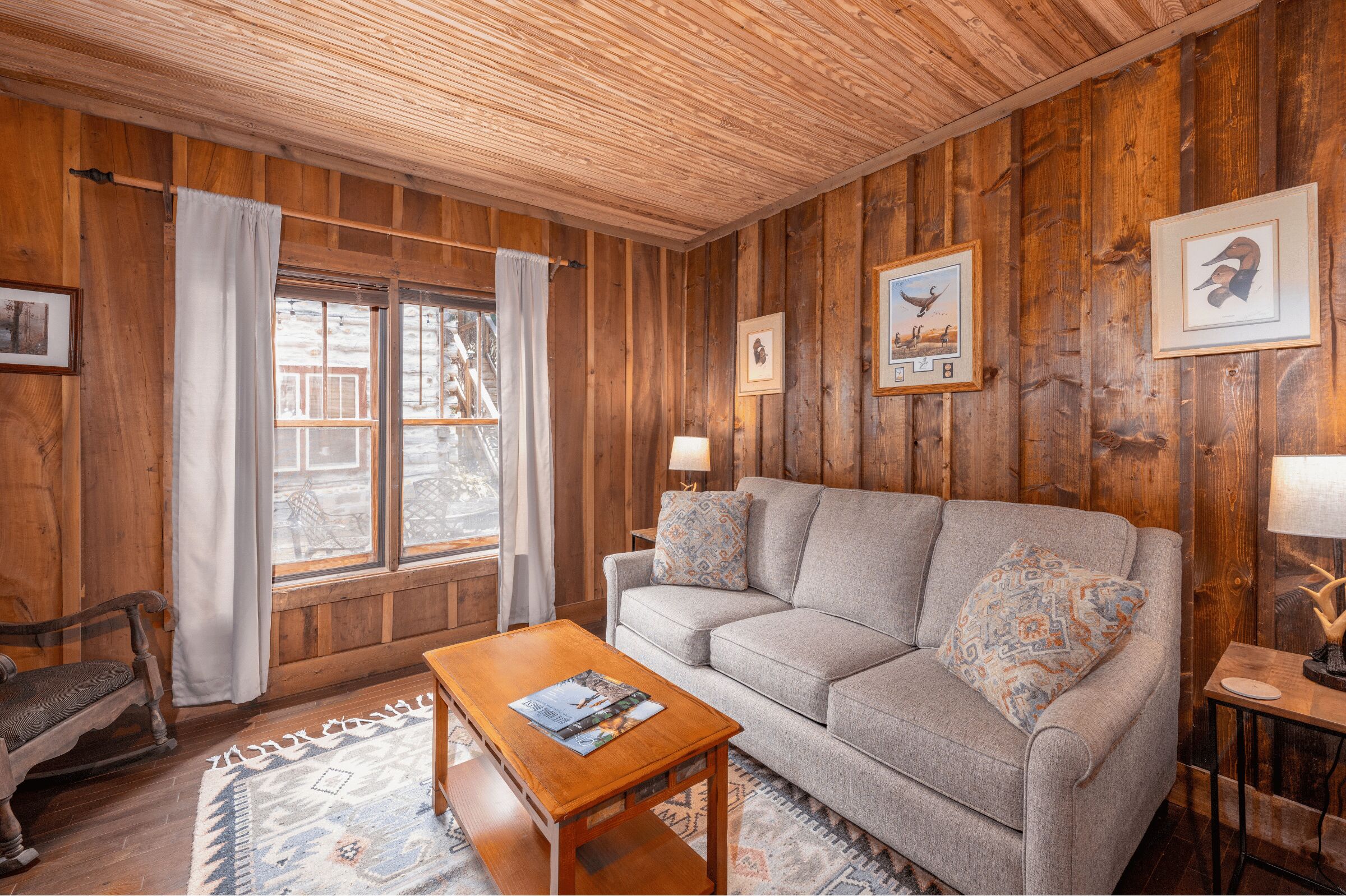 A wooden-paneled room with a sofa, a coffee table, and a rocking chair. There are framed pictures on the walls and a window with curtains.