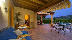 A covered patio with a blue sofa, a dining table, and chairs. There is a view of a mountain and a hedge in the background. - Ca'l Gall - Two Bedroom Villa, Sleeps 4 (Crestatx)