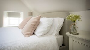 A neatly made bed with white and pink pillows, a vase with flowers on a bedside table, and a window providing natural light. - Eider Cottage (Buckie)