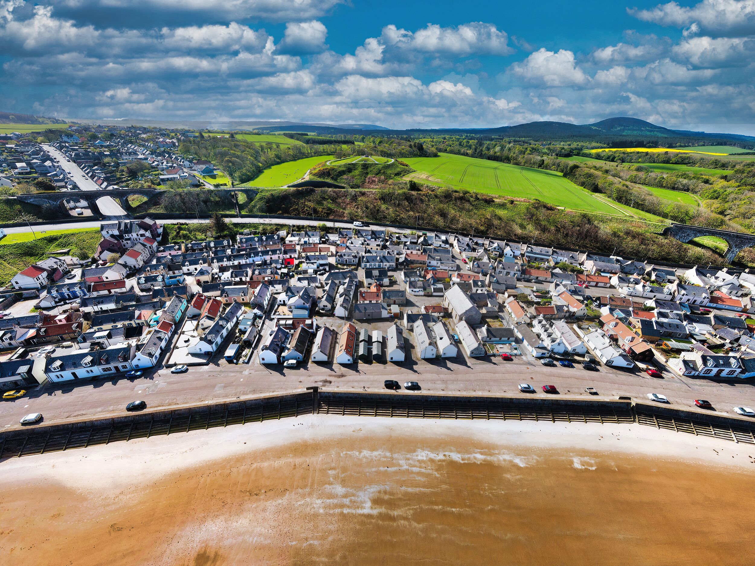 An aerial view of a coastal town with a sandy beach, a dense cluster of houses, and green fields in the background.