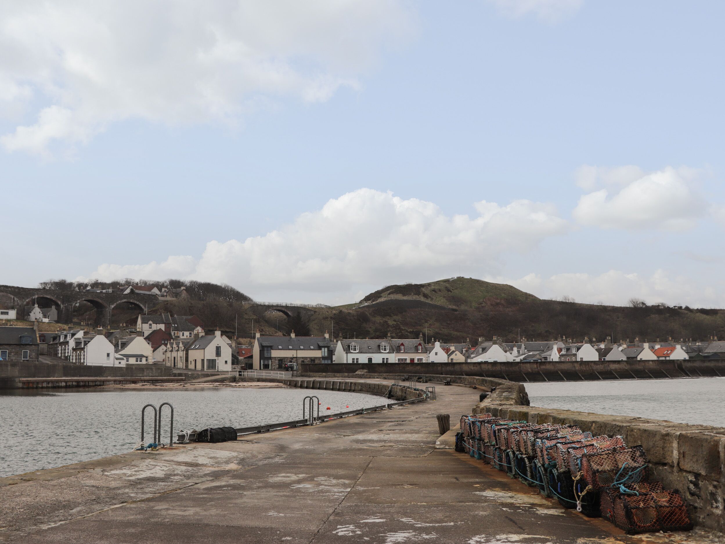 A coastal town with a pier leading to a harbor, houses along the waterfront, and a hill with a bridge in the background.