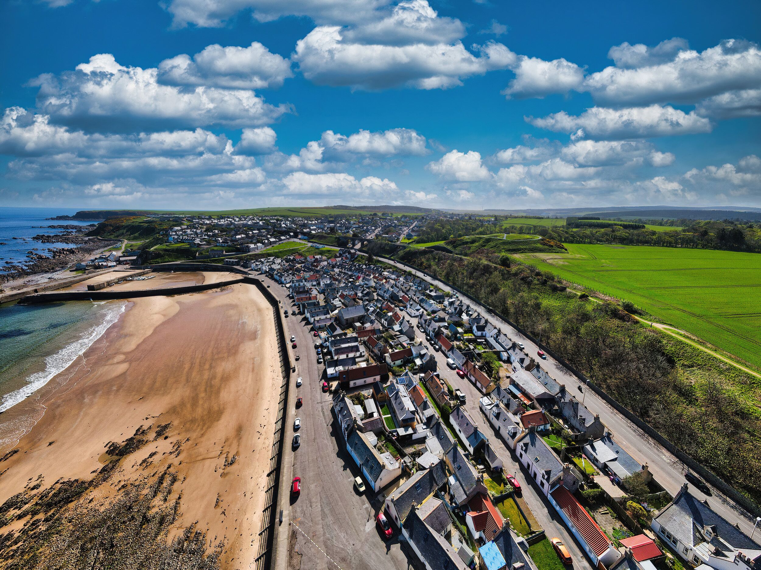 Vue aérienne d’une ville côtière avec une plage de sable, des immeubles résidentiels et des champs verdoyants.