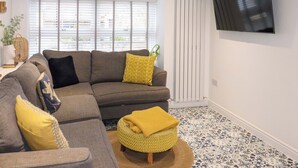 A modern living room with a grey sectional sofa, yellow cushions, a yellow knitted pouf, and a large flat-screen TV on the wall. - Eider Cottage (Buckie)
