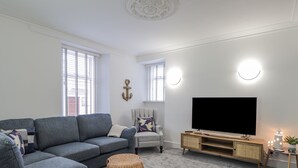 Una sala de estar con un sofá seccional gris, un pequeño taburete redondo, un televisor sobre un mueble de madera y una lámpara fijada a la pared.