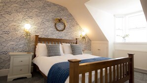 Un dormitorio con cama de madera, mesitas de noche blancas, papel tapiz estampado y techo inclinado.