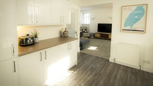 Una cocina moderna con armarios blancos, encimera de madera y un tostador. Hay una planta en la encimera y una cesta con objetos. Se ve un área de estar con un televisor y un sofá al fondo.