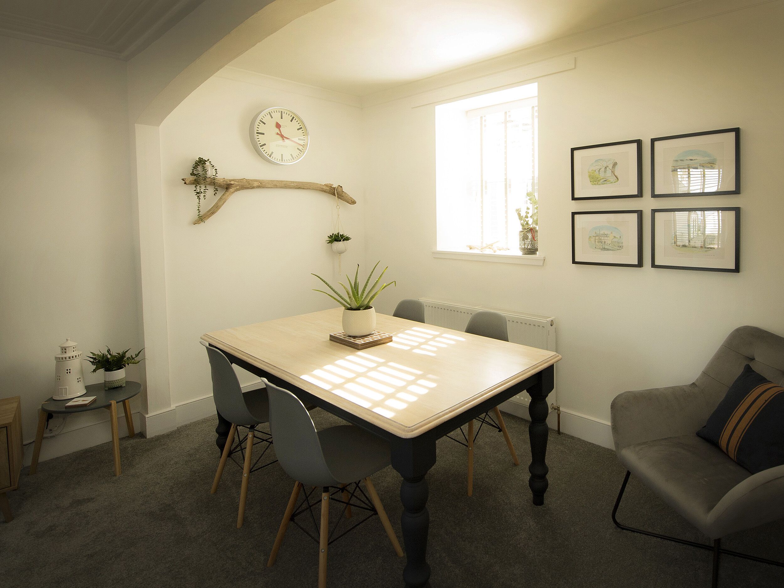 Une salle à manger avec une table en bois, quatre chaises, une plante en pot, une horloge murale et des tableaux encadrés.
