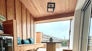 Eine Holzterrasse mit einem runden Tisch, Bänken, einer kleinen Hängeleuchte und Blick ins Freie.