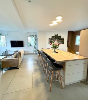 A modern kitchen with a wooden island, bar stools, and a dining table. There is a flat-screen TV mounted on the wall, a window with curtains, and a painting of a bridge.