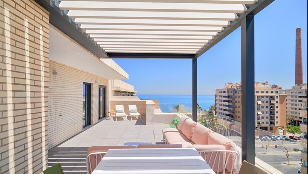 Un balcón moderno al aire libre con vista al mar, que incluye una zona de estar con sofá y sillas, y una barandilla de vidrio.