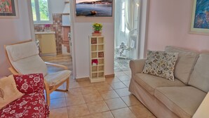 Een woonkamer met een beige bank, een rode fauteuil met bloemenprint, en een houten stoel. Een televisie is aan de muur bevestigd en toont een mooi landschap. Een klein kastje met decoratieve objecten en een bloevas is zichtbaar.