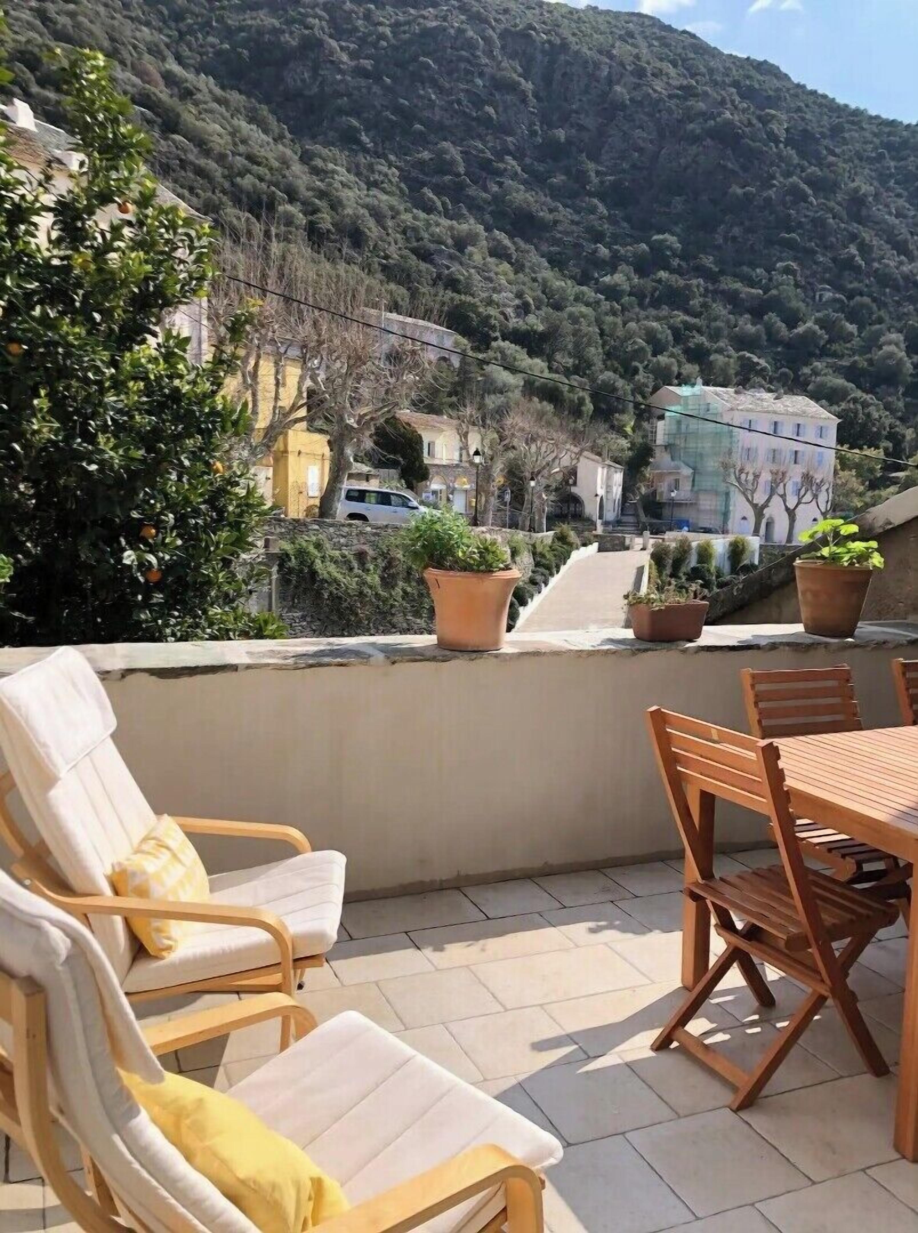 A terrace with outdoor furniture, potted plants, and a view of a hillside with buildings.
