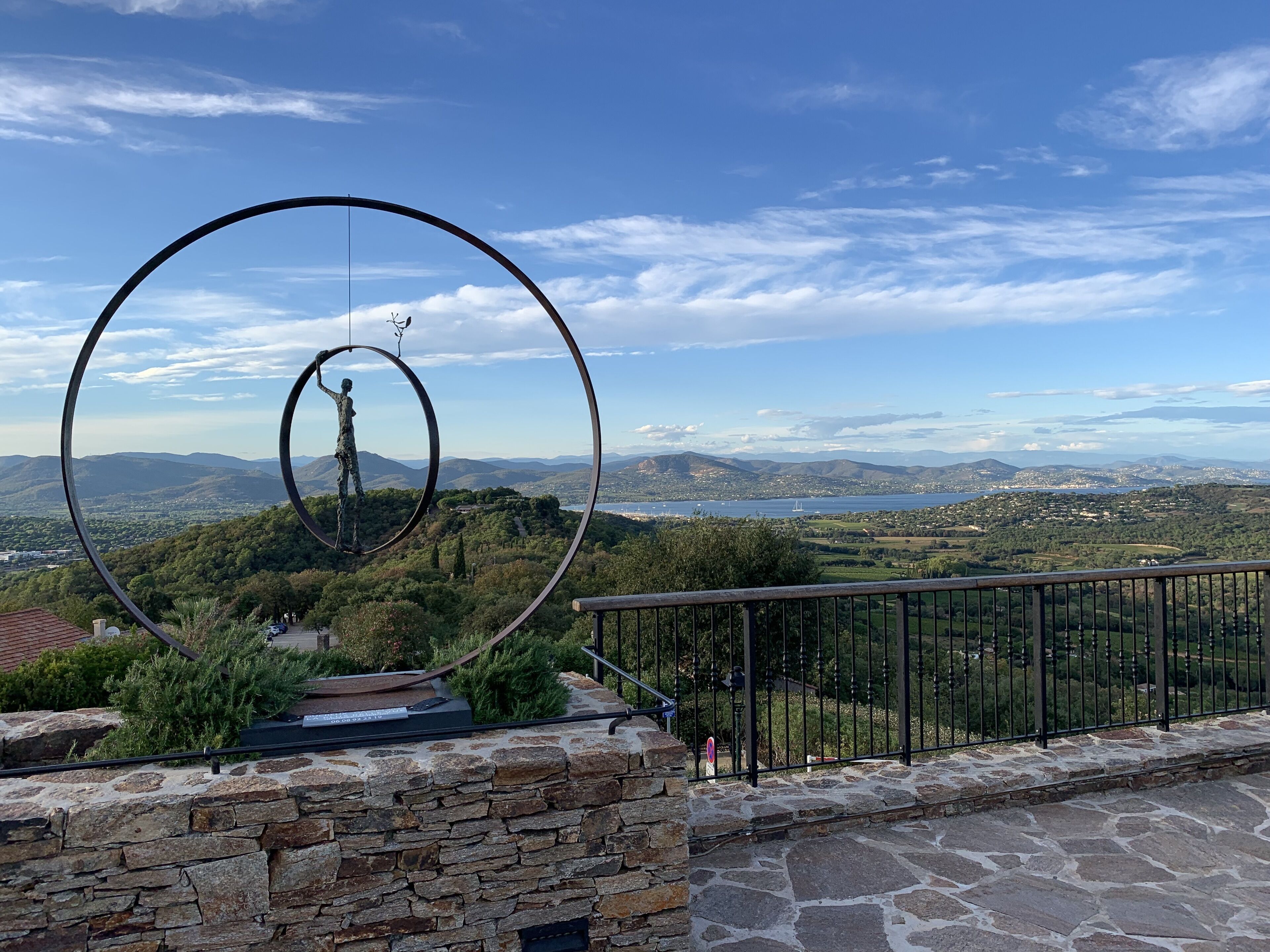 A stone wall with a metal sculpture of a figure holding a branch, overlooking a scenic landscape with mountains and a body of water.