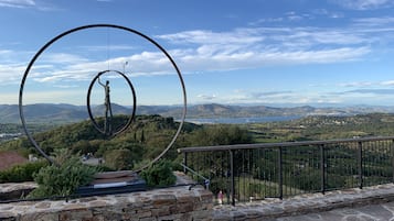 Un mur de pierre surmonté d’une sculpture métallique représentant une figure tenant une branche, dominant un paysage pittoresque avec des montagnes et une étendue d’eau.