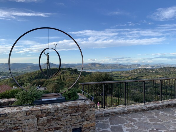 A stone wall with a metal sculpture of a figure holding a branch, overlooking a scenic landscape with mountains and a body of water.