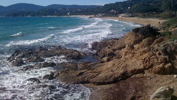 Paisagem costeira com terreno rochoso, uma praia arenosa e céu azul límpido.