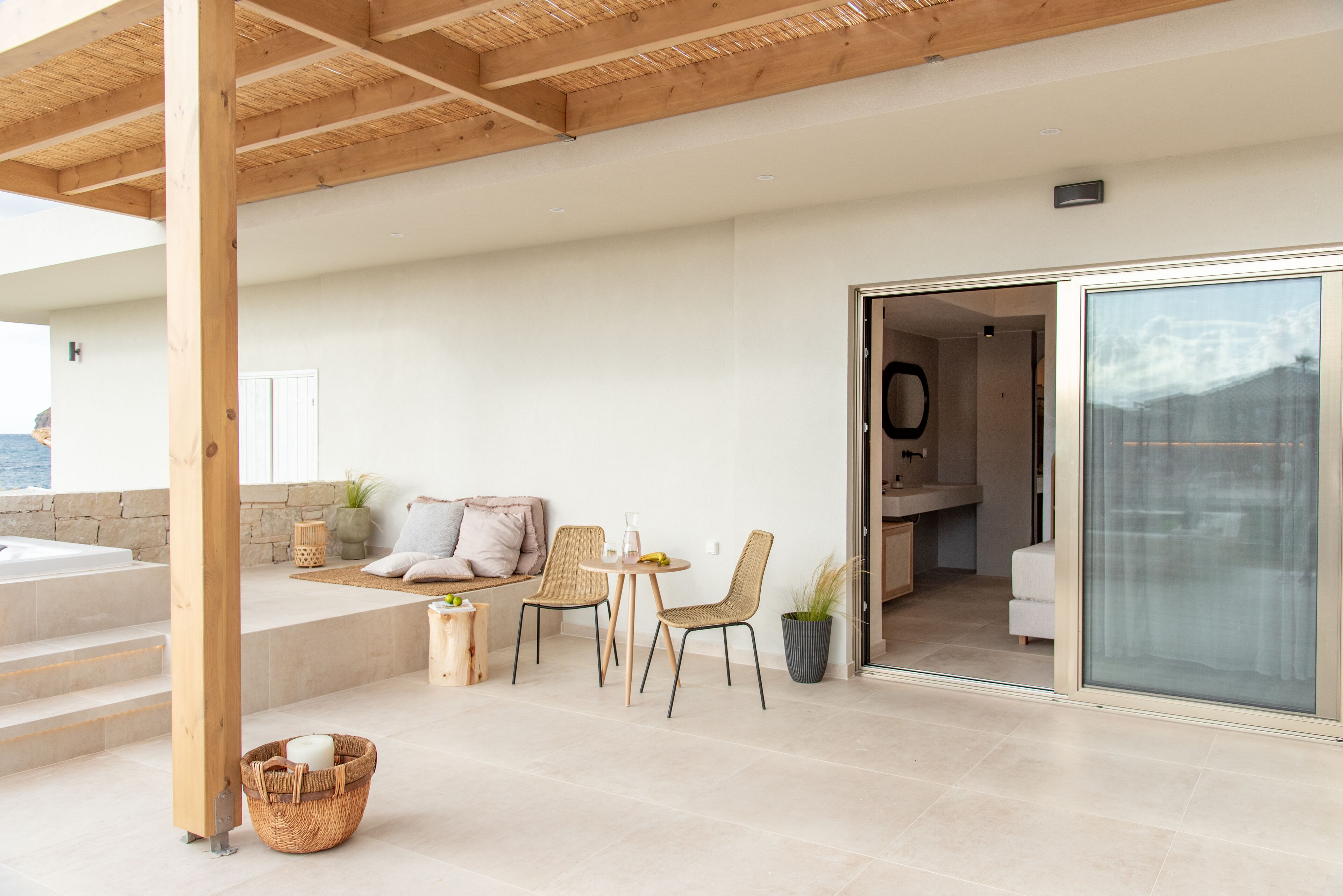 Uma área de estar moderna ao ar livre com pérgola de madeira, um espaço de convívio com cadeiras e mesa, um cesto com vela, e vista para o mar através de uma porta de vidro que dá para um banheiro.