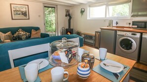 A cozy living room with a sofa, a coffee table with cups and a bread basket, a kitchen area with a washing machine, and a fireplace. - Chydour (Penzance)