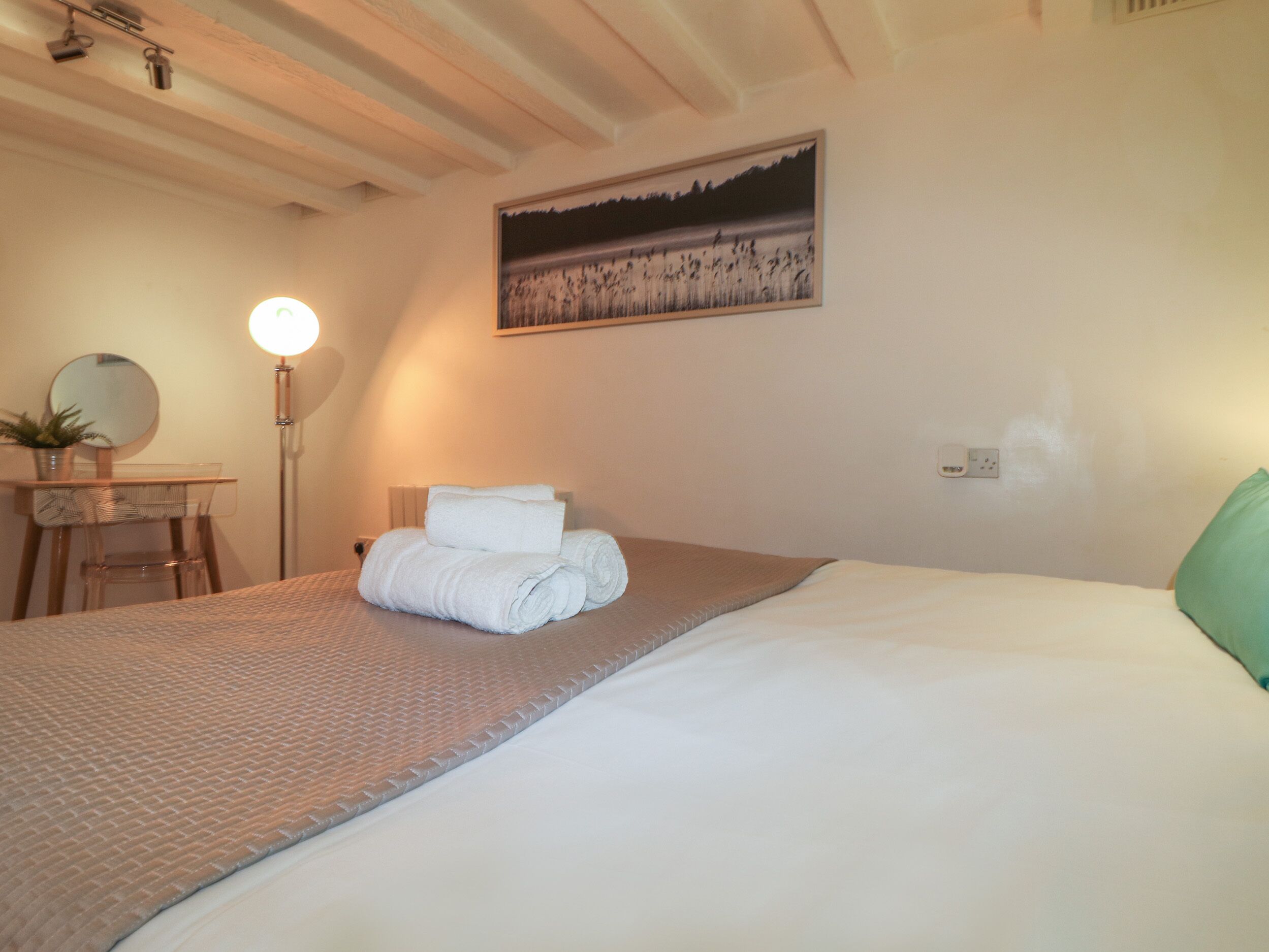 A neatly made bed with a rolled white towel, a bedside table with a mirror and a small plant, a floor lamp, and a painting on the wall.