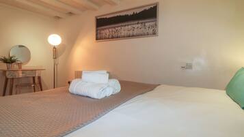 A neatly made bed with a rolled white towel, a bedside table with a mirror and a small plant, a floor lamp, and a painting on the wall.