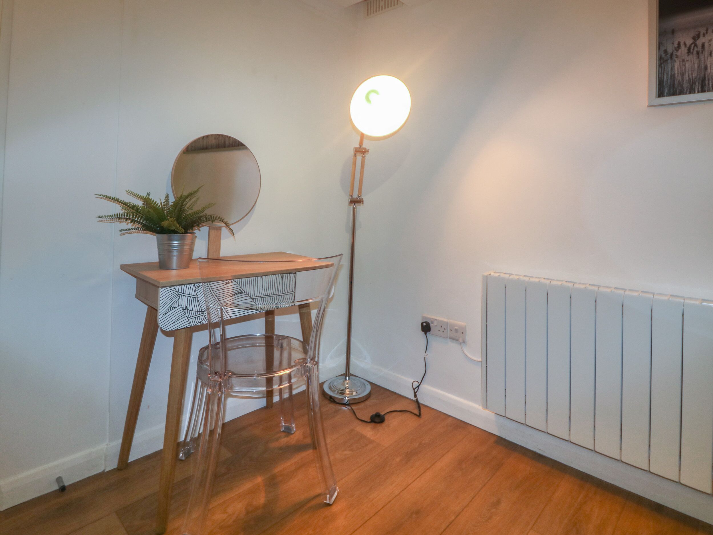 Una habitación con piso de madera, un escritorio con una silla transparente, una planta en maceta, un espejo redondo y una lámpara de pie.