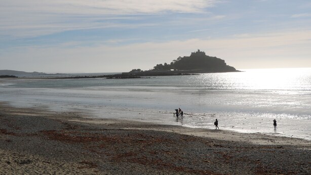 A beach with a castle-like structure in the distance, a clear sky, and a few people walking along the shore. - Chydour (Penzance)